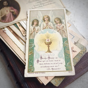 Puede incluir: Tarjeta religiosa antigua con tres ángeles, un cáliz y el texto "SAINTS ANGES DE DIEU". La tarjeta tiene una combinación de colores dorados, verdes y blancos. Se ven otras tarjetas y un libro encuadernado en cuero en el fondo.