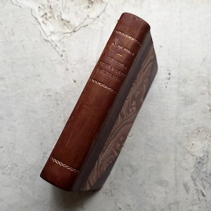 以下が含まれることがあります： 茶色の革の背表紙と金色の装飾が施されたヴィンテージ本。タイトルと著者は金色の文字でエンボス加工されています。表紙は茶色とクリーム色の模様です。クラシックな本です。
