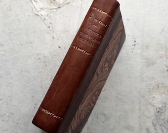 Op de afbeelding: Een vintage boek met een bruine leren rug en gouden details. De titel en auteur zijn in goud geëmbosseerd. De omslag heeft een patroon in bruin- en crèmetinten. Een klassiek boek.