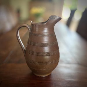 May include: A brown ceramic pitcher with a curved handle and a spout. The pitcher has horizontal bands of textured detailing around its body. The pitcher is sitting on a wooden surface.
