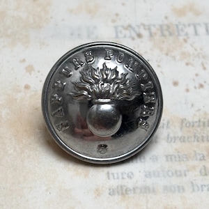 Può includere: Bottone color argento con testo in rilievo e un disegno centrale di un'urna in fiamme. Il bordo del bottone è leggermente rialzato e il testo è disposto a cerchio attorno all'immagine centrale. Lo sfondo è carta vintage.