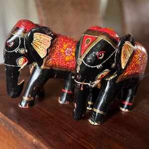 Könnte beinhalten: Zwei dekorative Holzfiguren in Form von Elefanten. Die Elefanten sind schwarz mit roten und goldenen Akzenten bemalt. Sie haben detaillierte Muster auf Rücken und Kopf und stehen auf einer Holzoberfläche.