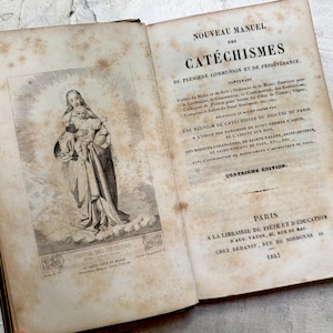 Puede incluir: Un libro antiguo abierto con páginas envejecidas y amarillentas. Una detallada ilustración en blanco y negro de una figura religiosa sosteniendo a un niño está en la página izquierda. Texto en francés impreso en la página derecha, con el título "Nouveau Manuel des Catéchismes."