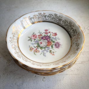 May include: A stack of white porcelain dishes with gold rims and floral center designs. The floral arrangement features pink, purple, and yellow flowers. The rims have a decorative gold pattern.