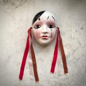 May include: A ceramic mask with a black and white design, red lips, and black eye details. The mask has red, brown, and white ribbons attached to the sides. The mask is mounted on a textured, off-white surface.