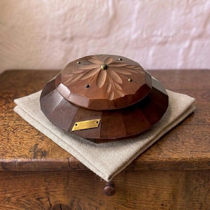 May include: A dark brown wooden lidded box with a faceted, multi-sided shape. The lid features a carved floral pattern with small metal studs. A rectangular brass-colored metal plate is attached to the side of the base.