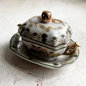 Può includere: Un piccolo piatto antico in porcellana con coperchio, appoggiato su un piattino abbinato. Il piatto e il coperchio sono decorati con motivi floreali nei toni del rosa, verde e oro. Il coperchio presenta un pomello dorato e i bordi sono rifiniti in oro.