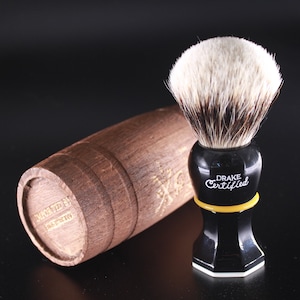 May include: A black and yellow shaving brush with a wooden barrel stand. The brush has a white and black badger hair brush head and the text "Drake Certified" on the handle.