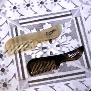 Pode incluir: Dois pentes, um bege claro e outro preto, com a palavra "Darwin" impressa na pega. Os pentes têm dentes muito juntos e uma pega curva. Estão sobre uma superfície estampada.