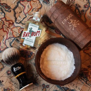Puede incluir: Un juego de afeitado con aftershave Drake's Bay Rum, una brocha de afeitar, un cuenco de madera con crema de afeitar y un barril de madera. La botella de aftershave tiene una etiqueta con texto. La brocha de afeitar tiene un mango negro con el texto "Drake Certified".