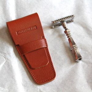 May include: A brown leather case with the word "DARWIN" embossed on the front. The case is closed and has a flap with a snap closure. A silver safety razor with a chrome finish is next to the case.