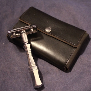 May include: A silver safety razor with a black leather case. The razor has a long, slender handle and a chrome blade.