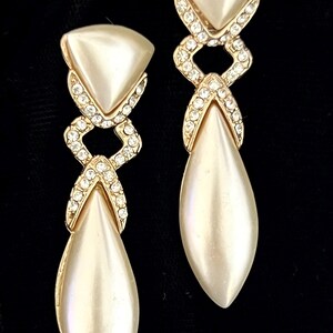 May include: A pair of elegant drop earrings with a pearlescent finish. Each earring features a triangular top, a diamond-shaped center adorned with small crystals, and a teardrop-shaped bottom. The earrings have a gold-tone metal setting.