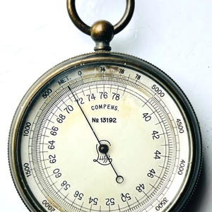 May include: A vintage brass altimeter with a white face and black markings. The altimeter has a needle that points to the number 50. The altimeter is labeled "COMPENS. No 13192".