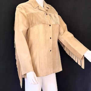 May include: A tan suede jacket with fringe detailing on the front and sleeves. The jacket has a button-up closure and a collared neckline.