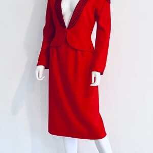May include: A red skirt suit with a fitted jacket featuring a black and red sequined trim on the collar and shoulders. The skirt is knee-length and has a straight silhouette.