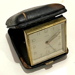 May include: A vintage gold-toned travel alarm clock with a white face and black hands. The clock is in a black leather case with a worn, distressed look. The clock face reads "Sema Seven Jewels".