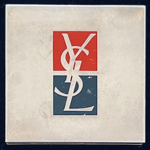 May include: Square, silver-toned compact with a red and blue logo. The logo features the letters "YSL" in a stylized, white font. The compact has a brushed metal finish and a simple, elegant design.