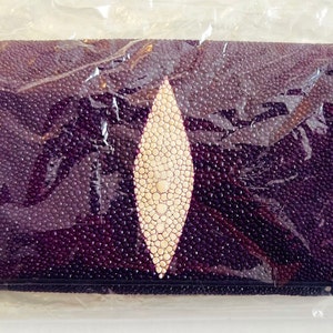 May include: A rectangular, dark purple wallet with a textured surface and a central, light-colored, oval-shaped design. The wallet is wrapped in clear plastic.