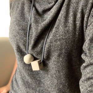 Può includere: Una collana con un ciondolo a forma di cubo in cemento grigio e una sfera rotonda su un cordoncino grigio. La collana è indossata da una persona che indossa un maglione grigio.