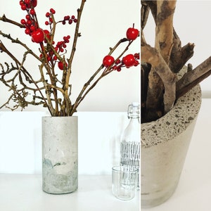 Puede incluir: Un jarrón cilíndrico de hormigón contiene ramas con bayas y manzanas rojas. El jarrón es de color gris claro con un aspecto moteado. Un vaso transparente y una botella decorativa están en el fondo. Tema: decoración del hogar.