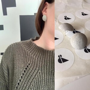 Può includere: Un paio di orecchini rotondi in cemento grigio. Gli orecchini sono mostrati sull'orecchio di una persona e su uno sfondo bianco con adesivi.