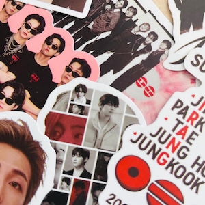 May include: A collection of die-cut stickers featuring various photographic portraits of a group of young men, some wearing sunglasses, alongside a white sticker with red circular icons and the text JIN PARK TAE JUNG HO JUNGKOOK 2026. 03.