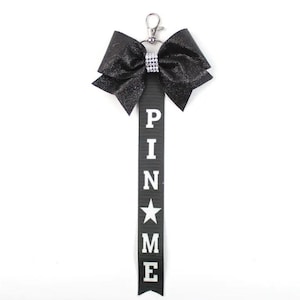 May include: Black sparkly bow with a silver rhinestone centre attached to a black ribbon with the word "PIN ME" in white letters and a white star.