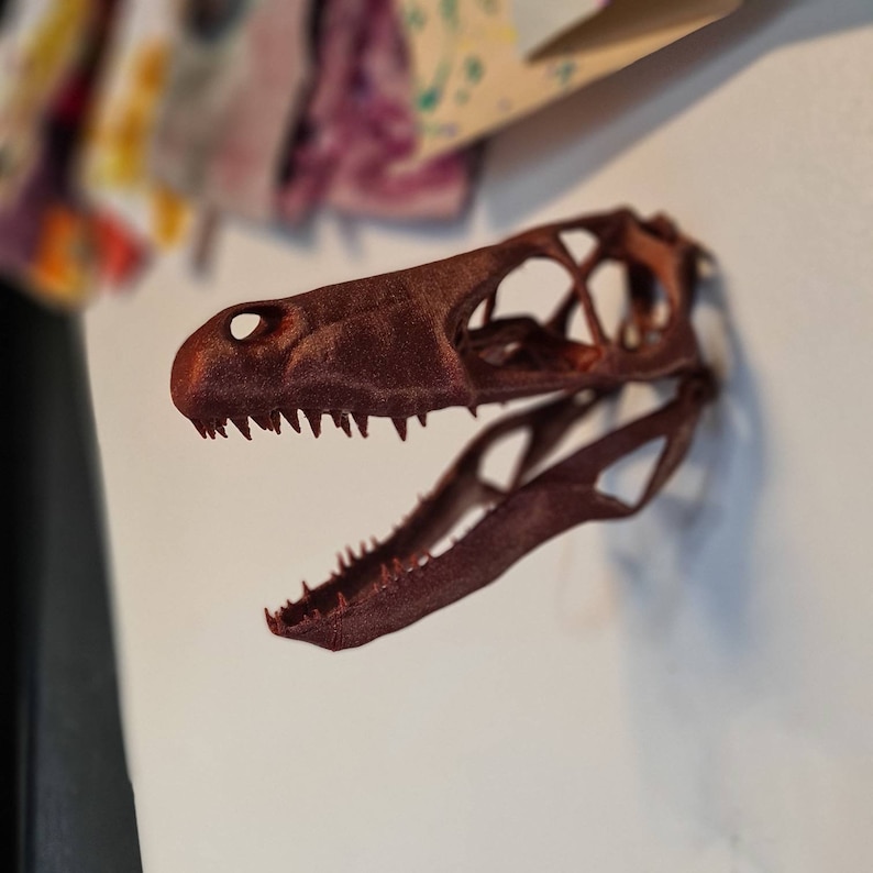 May include: A 3D printed dinosaur skull model in a rusty brown color. The skull is open-mouthed and has sharp teeth. The model is mounted on a white wall.