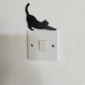 Könnte beinhalten: Schwarze Metallkatzensilhouette, die sich über einem weißen Lichtschalter ausdehnt. Die Katze befindet sich in einer spielerischen Pose und verleiht der Wand eine dekorative Note. Der Lichtschalter ist rechteckig mit einem kleinen Schalter.
