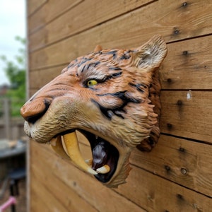 Könnte beinhalten: Nahaufnahme einer knurrenden Säbelzahntiger-Kopfskulptur. Die Skulptur ist aus bemaltem Harz gefertigt und weist realistische Details auf, darunter scharfe Zähne und ein strukturiertes Fellmuster.