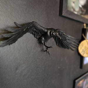 May include: A matte black wall-mounted sculpture of a raven in flight with wings fully extended. The bird is depicted with detailed feathers, a sharp beak, and talons reaching downward, creating a dynamic silhouette against the dark surface.