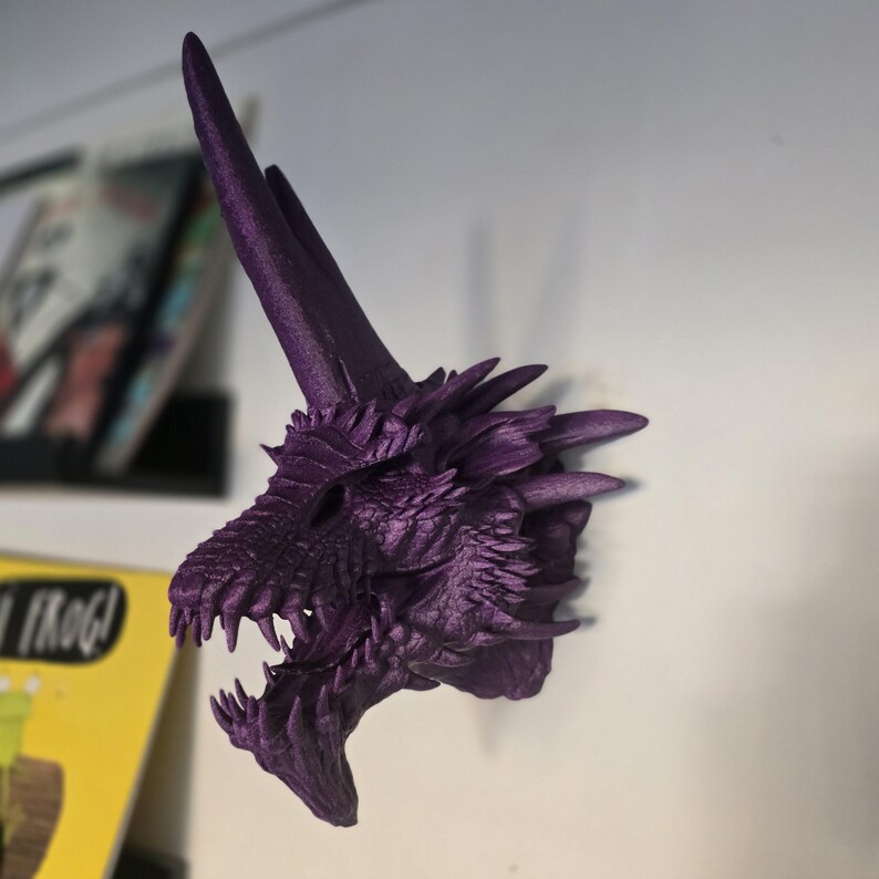 May include: Purple 3D printed dragon head wall mount. The dragon has a long, pointed horn and an open mouth with sharp teeth.