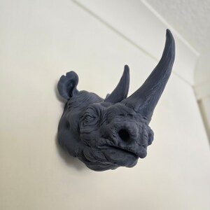 Peut inclure: Une sculpture de t&ecirc;te de rhinoc&eacute;ros gris fonc&eacute; mont&eacute;e sur un mur blanc. La sculpture est d&eacute;taill&eacute;e avec une peau textur&eacute;e et une corne pro&eacute;minente.