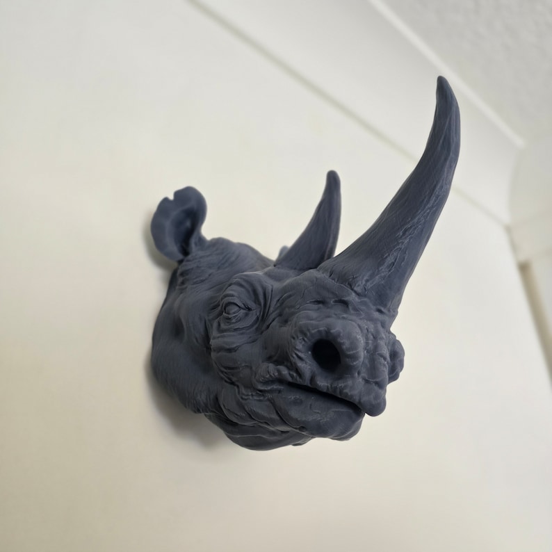 Peut inclure: Une sculpture de t&ecirc;te de rhinoc&eacute;ros gris fonc&eacute; mont&eacute;e sur un mur blanc. La sculpture est d&eacute;taill&eacute;e avec une peau textur&eacute;e et une corne pro&eacute;minente.