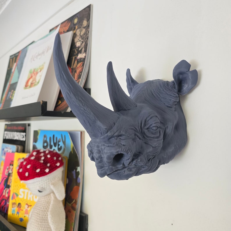 Peut inclure: Une t&ecirc;te de rhinoc&eacute;ros grise en guise de d&eacute;coration murale. La t&ecirc;te est tourn&eacute;e vers la droite de l'image. Le rhinoc&eacute;ros a une grande corne et une surface textur&eacute;e.