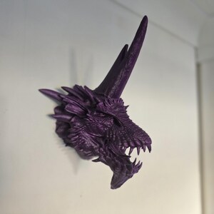 May include: Purple 3D printed dragon head wall mount. The dragon has a long, pointed horn and an open mouth with sharp teeth.