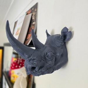 Peut inclure: Une t&ecirc;te de rhinoc&eacute;ros grise en guise de d&eacute;coration murale. La t&ecirc;te est tourn&eacute;e vers la droite de l'image. Le rhinoc&eacute;ros a une surface textur&eacute;e et un visage d&eacute;taill&eacute;.