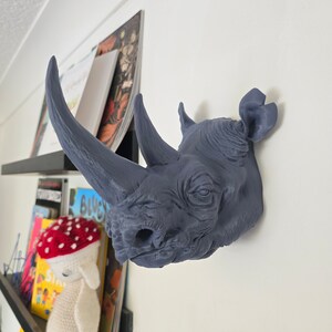 Peut inclure: Une t&ecirc;te de rhinoc&eacute;ros grise mont&eacute;e sur un mur blanc. Le rhinoc&eacute;ros a une grande corne et une surface textur&eacute;e.