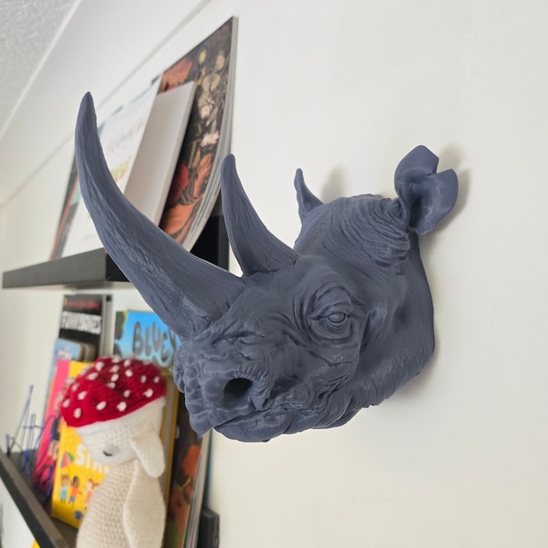 Peut inclure: Une t&ecirc;te de rhinoc&eacute;ros grise mont&eacute;e sur un mur blanc. Le rhinoc&eacute;ros a une grande corne et une surface textur&eacute;e.