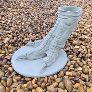 May include: A grey, 3D printed dinosaur foot planter. The planter is shaped like a dinosaur foot with three toes and a textured surface. The planter is sitting on a bed of small, grey stones.
