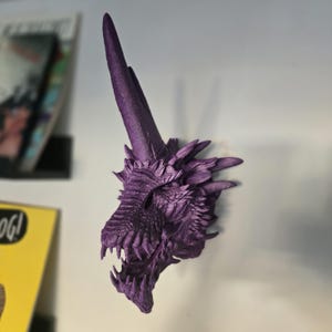 May include: A purple 3D printed dragon head wall mount with a long, pointed horn. The dragon's mouth is open, revealing sharp teeth.