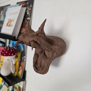 May include: A brown ceramic sculpture of a horned viper with its mouth open, showing its fangs. The sculpture is mounted on a white wall.