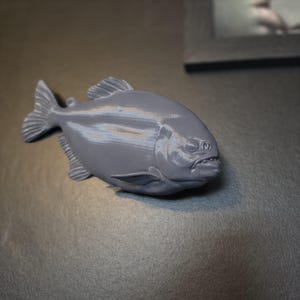 May include: A three-dimensional printed figurine of a piranha fish in a solid matte grey colour. The fish features a deep, rounded body, a prominent dorsal fin, a fan-shaped tail, and visible gill and mouth details rendered in a smooth plastic finish.
