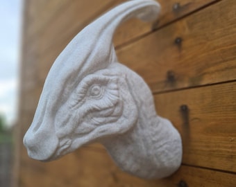 Op de afbeelding: Witte dinosauruskop sculptuur met een getextureerd oppervlak. Het beeld is gemonteerd op een houten muur.