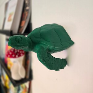 May include: A green plastic turtle figurine with a detailed shell and textured legs. The turtle is mounted on a white wall.