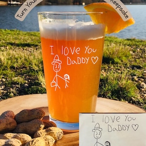 May include: A clear glass with a drawing of a stick figure family on it. The glass is filled with a light orange beverage and has a slice of orange on the rim. The drawing says "I love you Daddy" and is surrounded by peanuts on a wooden surface.