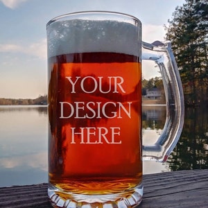 May include: A clear glass beer mug with a handle, filled with a dark amber beer and a white head. The mug is sitting on a wooden surface with the text "YOUR DESIGN HERE" etched on the side. The background is a blurry image of a lake and trees.