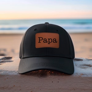 May include: Black baseball cap with a brown leather patch that says "Papa" on it. The cap is sitting on a sandy surface.
