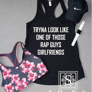 May include: Black tank top with the text "Tryna look like one of those rap guys girlfriends." A black sports bra with a pink floral pattern is next to the tank top. A black cap with a white Nike logo and a white smartwatch are also in the image.